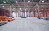 A large, well-lit, empty warehouse with polished concrete floors and stacks of grey and orange pallets in the midground.