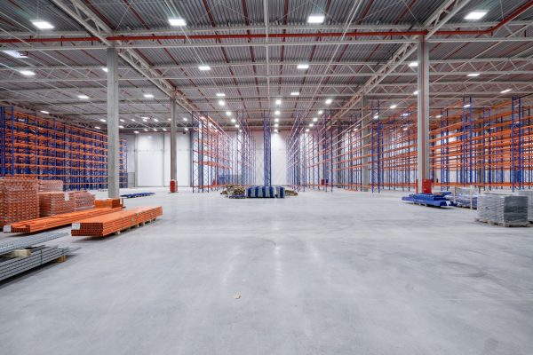 A large, well-lit, empty warehouse with polished concrete floors and stacks of grey and orange pallets in the midground.