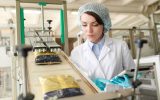 A young woman working at a modern food factory and looking at macaroni bags sliding down conveyor belt.