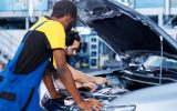 Two male auto repair technicians work together to diagnose and treat the engine of gray sedan with its hood open.