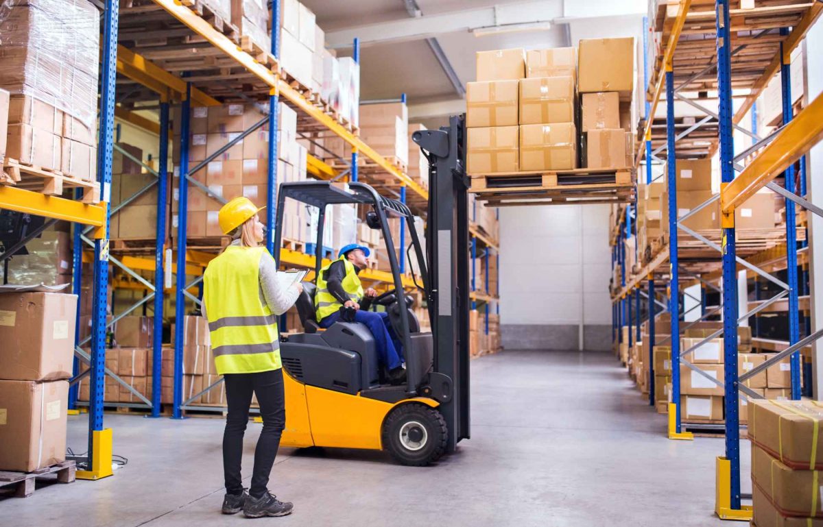 Forklift driver in visibility vest and hardhat raising pallet toward rack while supervisor in vest and hardhat observes