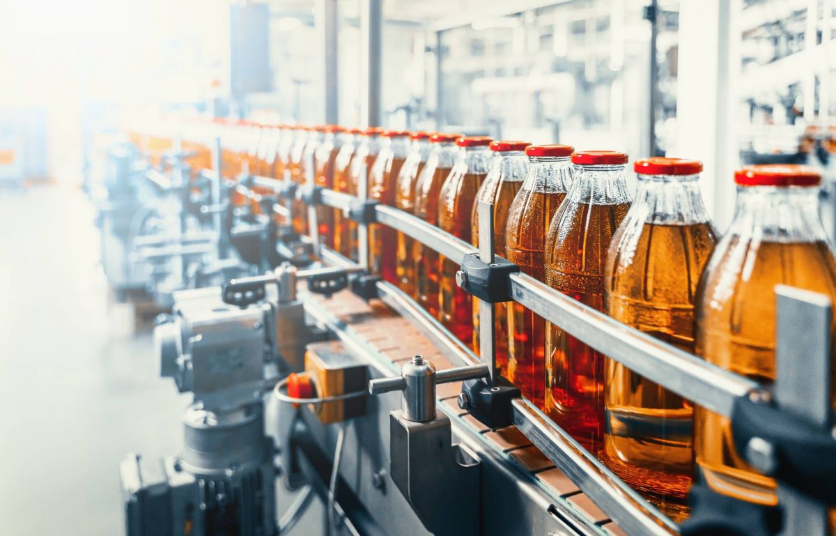 A large conveyor belt moves glass bottles filled with a brown liquid and red cap to a new location.