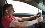 A man in a red shirt tightly gripping the steering wheel of his car and shouting as he seems to lose control of the vehicle.