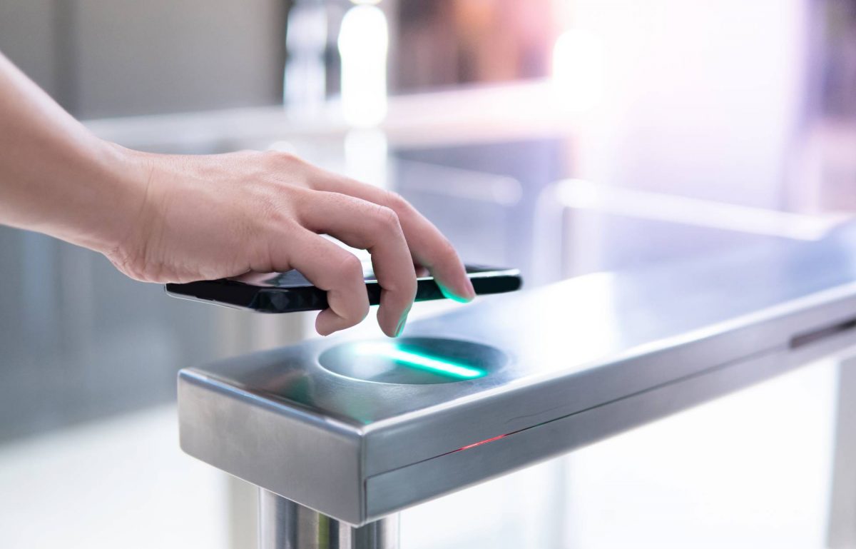 A person's hand holding a black smartphone over a green scanner built into a metal gate structure for access control.